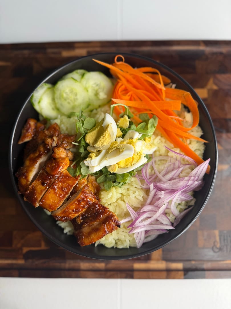 A black bowl of rice with sliced chicken, cucumbers, carrots, red onions, arugula, and hard-boiled eggs.